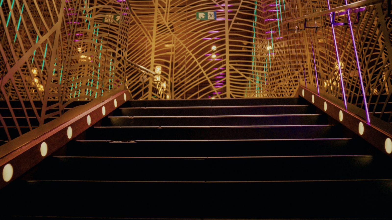 Escalera de acceso con balizas en los peldaños, envuelta por una celosía dorada de líneas curvas iluminada.