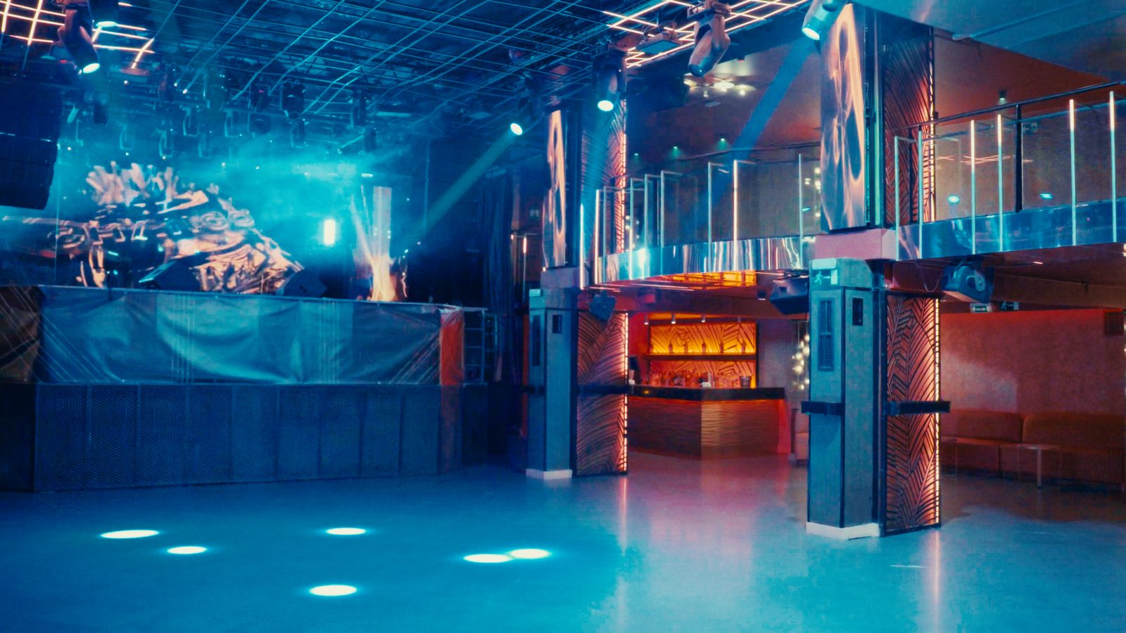 Cabina de DJ y frente de escenario bajo luz azul, con la entreplanta y la pista en primer plano.
