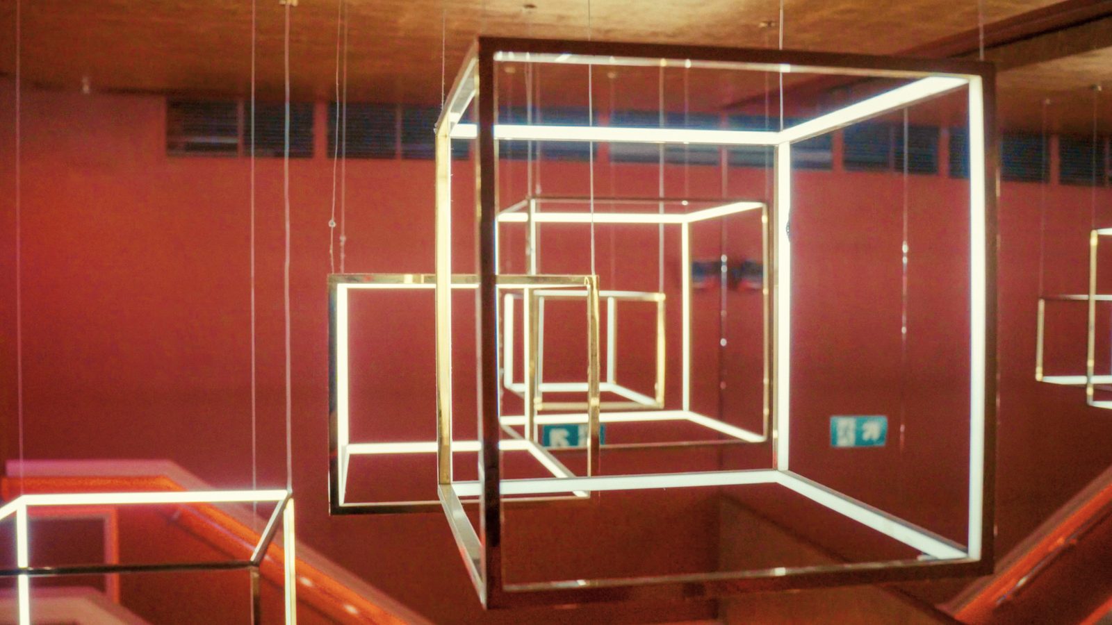 Estructuras cúbicas de tubos de luz blanca suspendidas en un espacio de paredes rojas.