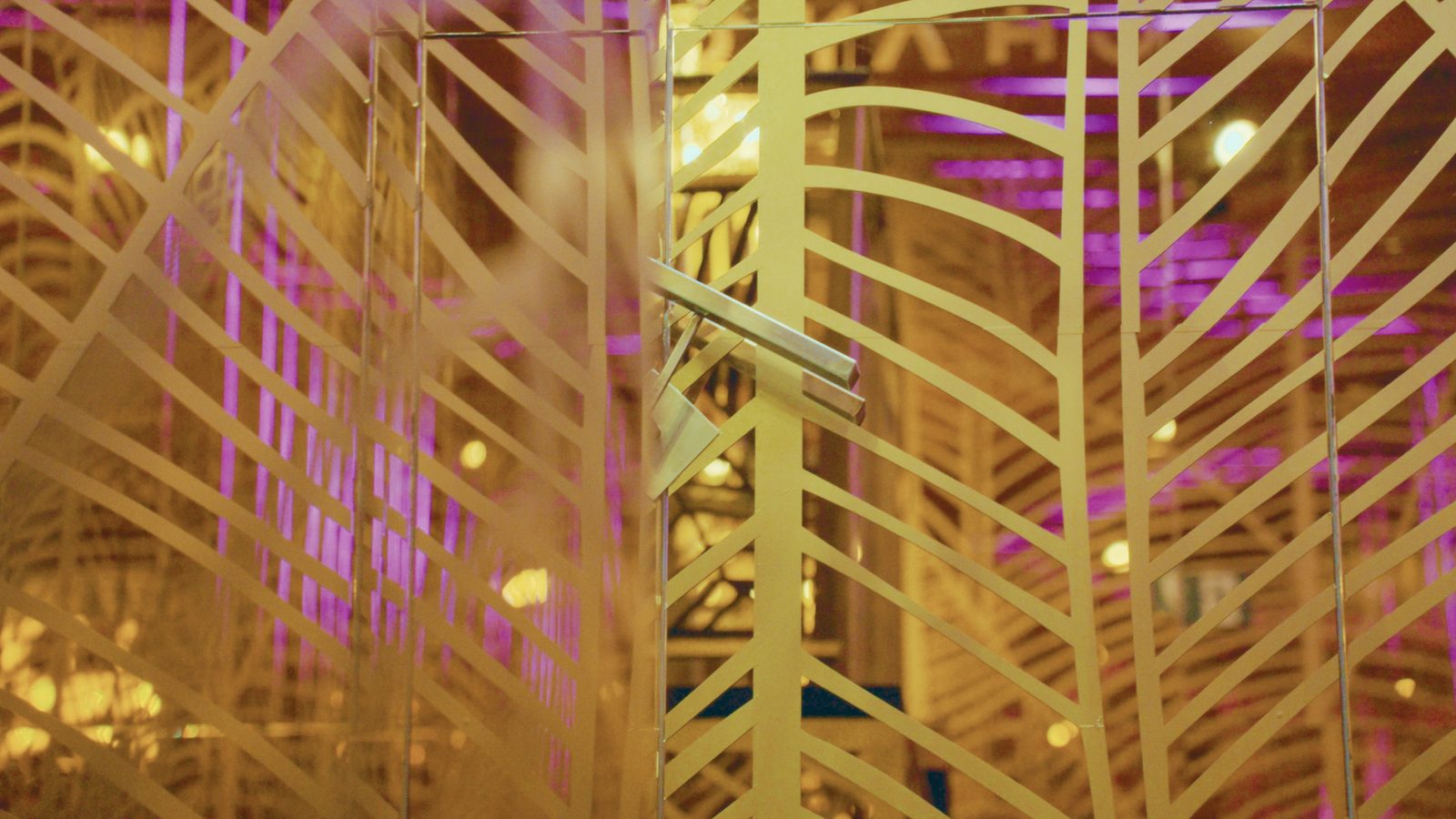 Detalle de la celosía dorada de hojas con reflejos de luz rosa y morada.